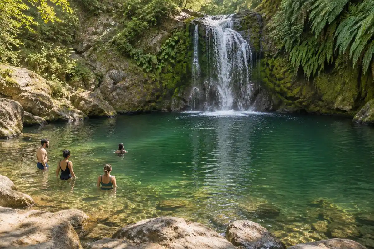 Wide swimming hole beneath a modest waterfall with people wading in the shallows
