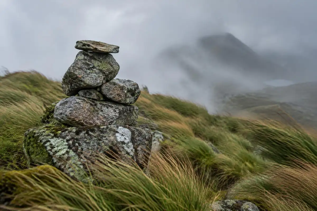 Weathered stone cairn with tussocky grass and blowing mist behind