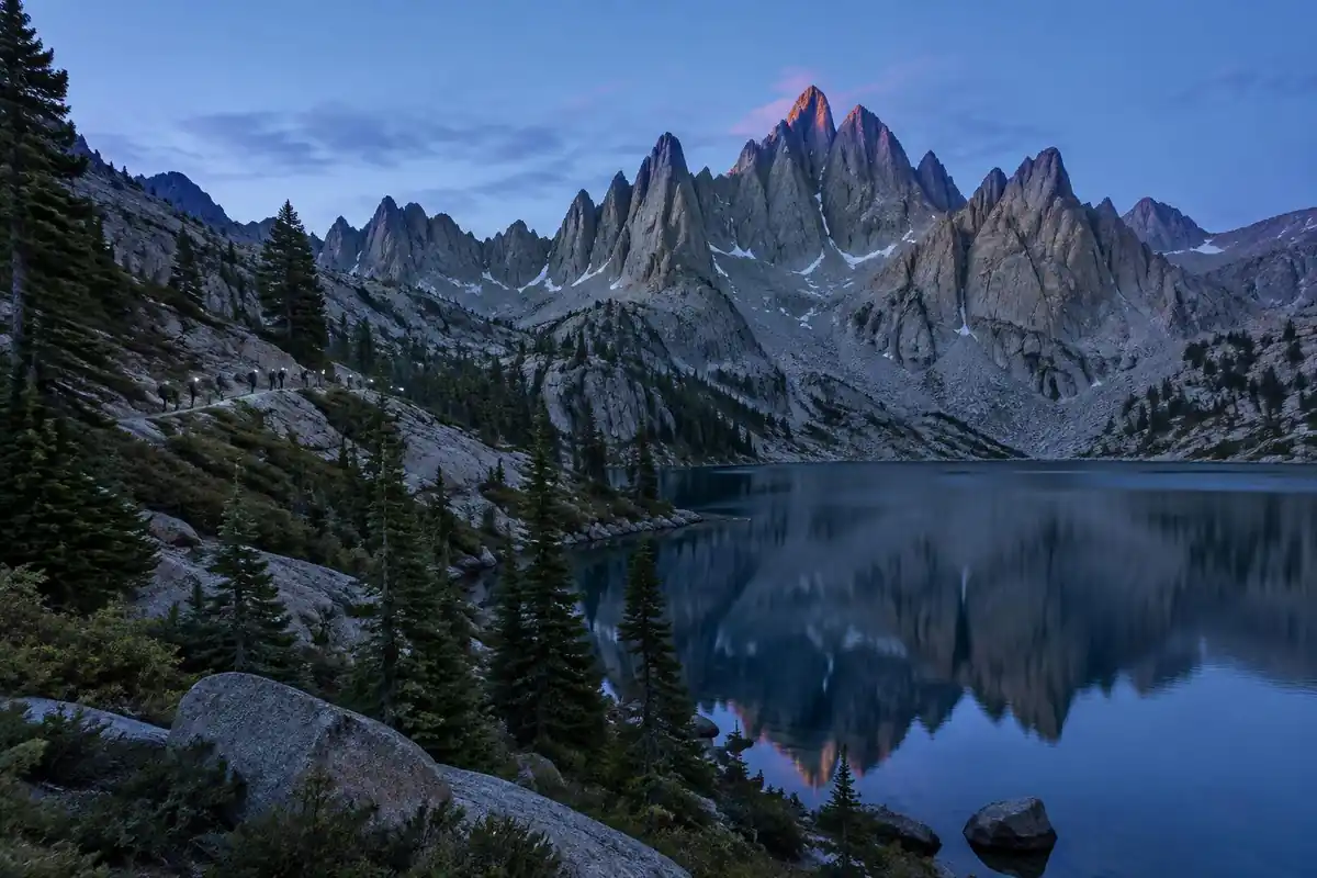 Alpine lake beneath jagged granite needles at first light, headlamps on a trail